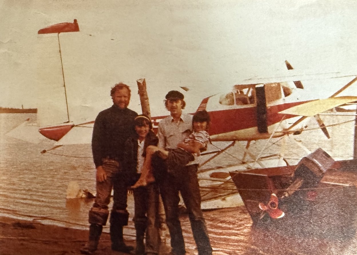 Vintage color photo: two men and two girls standing at the water's edge in front of a red and white floatplane.
